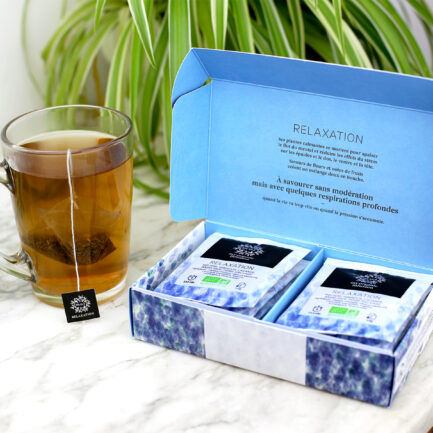 Coffret relaxation 12 infusettes ouvert sur un meuble en marbre avec une tasse qui infuse à sa gauche.