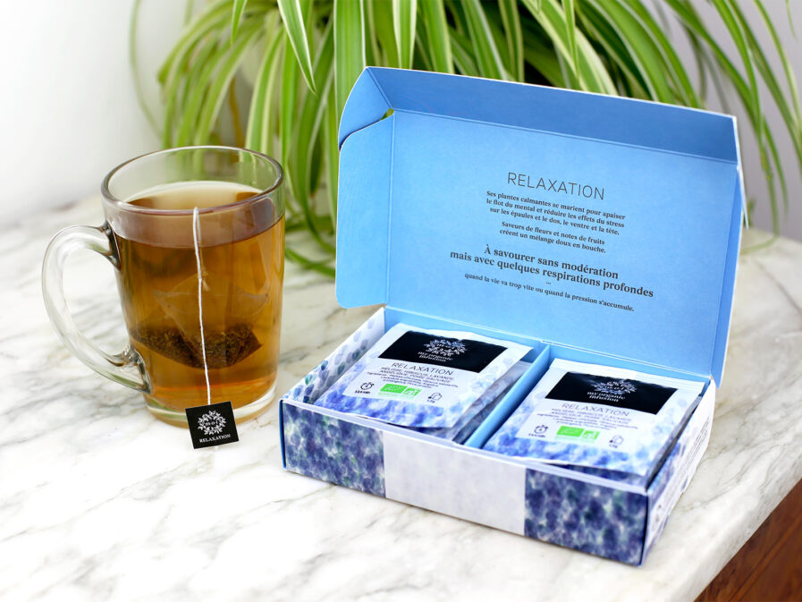 Coffret relaxation 12 infusettes ouvert sur un meuble en marbre avec une tasse qui infuse à sa gauche.